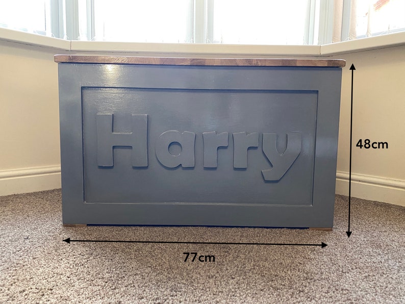 May include: A rectangular wooden toy chest painted in a matte gray finish with the name Harry in raised lettering on the front panel. The chest features a natural wood top lid and measures 77 cm in width by 48 cm in height as indicated by arrows.