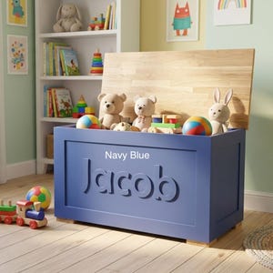 May include: A rectangular navy blue wooden toy chest with a natural wood-toned lid. The front of the chest features the name Jacob in large, raised, matching navy blue lettering. The chest is filled with various plush toys and colorful balls.