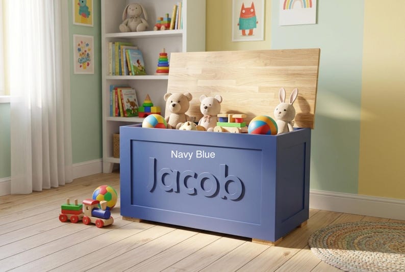 May include: A rectangular navy blue wooden toy chest with a natural wood-toned lid. The front of the chest features the name Jacob in large, raised, matching navy blue lettering. The chest is filled with various plush toys and colorful balls.