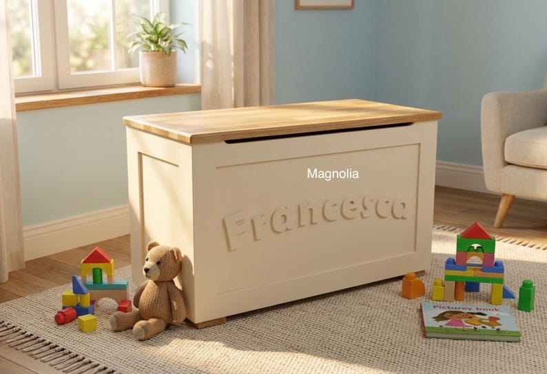 May include: A rectangular cream-colored wooden toy chest with a natural wood-toned top lid. The front panel features the name Francesca in raised lettering and the word Magnolia printed above it. The chest is surrounded by colorful building blocks.