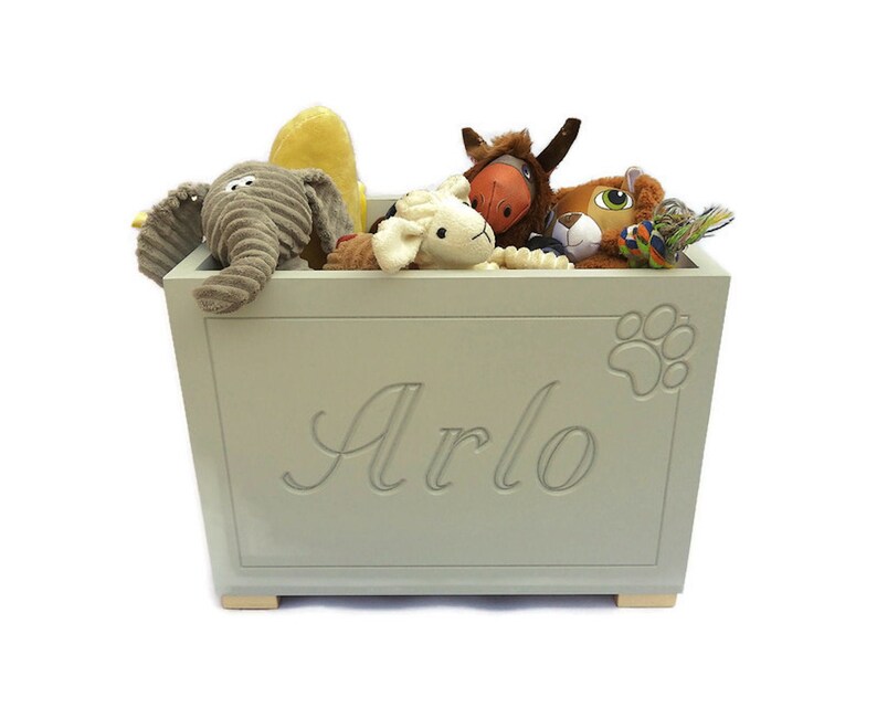Op de afbeelding: Een lichtgrijze rechthoekige houten speelgoedkist met de naam Arlo in cursief schrift gegraveerd op het voorpaneel, vergezeld van een reli&euml;f pootafdrukontwerp. De kist is gevuld met diverse pluche knuffeldieren, waaronder een olifant.