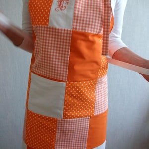 Peut inclure: Tablier en patchwork orange et blanc avec un carré blanc au centre orné d'une lettre "L" brodée.