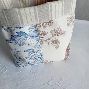 Peut inclure: Un panier de rangement en tissu avec un dessus replié. Le panier présente un motif patchwork avec des motifs floraux bleus et marron sur fond crème. Le bord supérieur a un motif rayé beige et blanc.