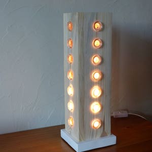 Peut inclure: Une lampe en bois avec une base blanche et une forme carrée. La lampe a une série de découpes circulaires sur les côtés, chacune avec une ampoule à l'intérieur. Les ampoules sont allumées, projetant une lueur chaude.