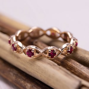 Può includere: Un anello color oro con un motivo ripetuto di piccole pietre preziose rosse e rotonde. L'anello è posto su uno sfondo di sottili bastoncini di legno marrone. Le pietre preziose sono equidistanti.