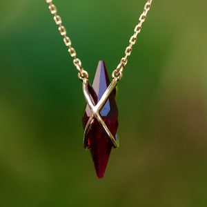 May include: A necklace featuring a deep red, faceted, diamond-shaped pendant suspended from a thin gold chain. The pendant is secured by two thin, crossed gold metal wires that wrap around the centre of the stone, creating a secure setting.