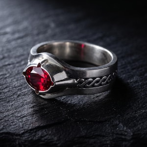 May include: A silver ring featuring a central oval-cut deep red gemstone held by four prongs. The band has a polished finish and includes a decorative braided or twisted metal pattern along the side, creating a textured look on the metal surface.