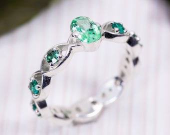 May include: A silver ring with an oval-cut green gemstone at the center and smaller round green gemstones set along the band. The ring has a twisted design and is set against a blurred background.