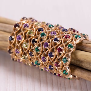 May include: A stack of gold rings with intricate designs and colourful gemstones. The rings are arranged on a wooden surface.