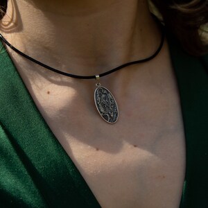 May include: Silver pendant necklace with an oval pendant featuring an engraved design. The pendant hangs from a black cord. The person is wearing a dark green blazer.