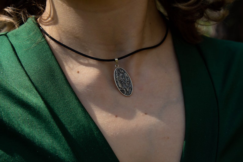 May include: Silver pendant necklace with an oval pendant featuring an engraved design. The pendant hangs from a black cord. The person is wearing a dark green blazer.