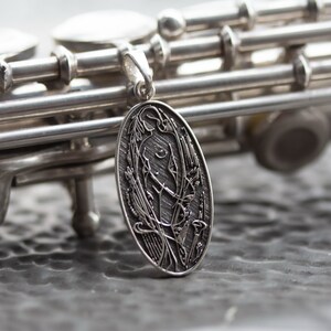 May include: A silver pendant with an oval shape, featuring an engraved design of a figure surrounded by floral elements. The pendant hangs in front of a silver flute, with the instrument's keys and body visible.