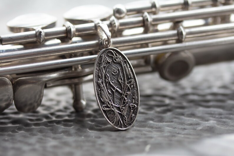May include: A silver pendant with an oval shape, featuring an engraved design of a figure surrounded by floral elements. The pendant hangs in front of a silver flute, with the instrument's keys and body visible.