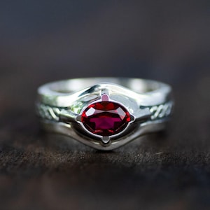 May include: A polished silver ring featuring a central marquise-cut deep red gemstone set in a smooth bezel. The band displays a subtle braided or woven texture along the sides, leading to a raised, contoured setting that holds the vibrant red stone.