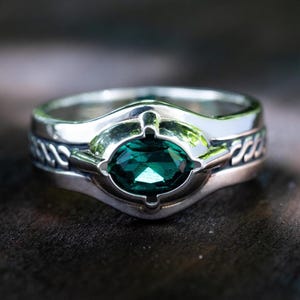 Op de afbeelding: Een zilveren ring met een grote, ovaal geslepen, smaragdgroene edelsteen in het midden. De ring heeft een gedetailleerd ontwerp met een gevlochten patroon op de band. De zetting heeft een contrasterende vorm.