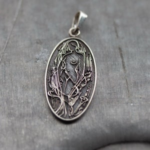 May include: Oval silver pendant with a detailed engraved design. The pendant features a stylized female figure with floral and abstract elements. The bail is a simple loop. The pendant is displayed on a gray surface.