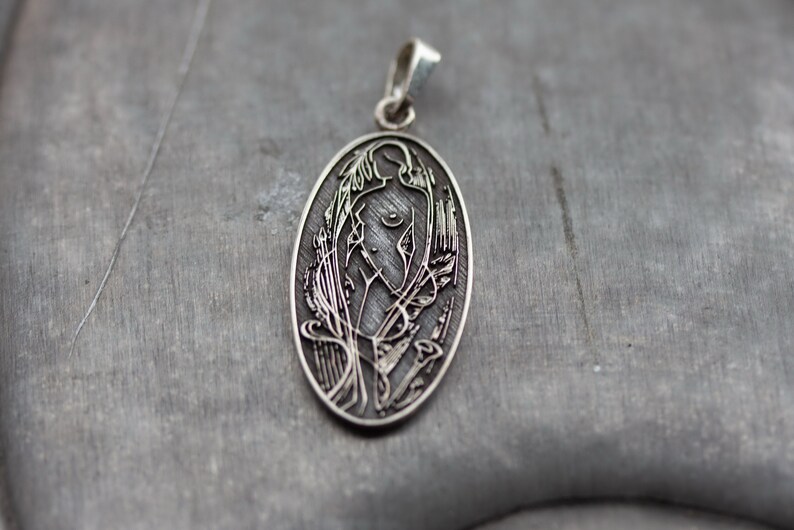 May include: Oval silver pendant with a detailed engraved design. The pendant features a stylized female figure with floral and abstract elements. The bail is a simple loop. The pendant is displayed on a gray surface.
