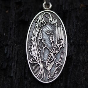 May include: Oval silver pendant featuring an engraved Art Nouveau style design. The pendant depicts a nude female figure amidst stylized floral and plant motifs. The pendant hangs from a silver chain against a dark, textured background.