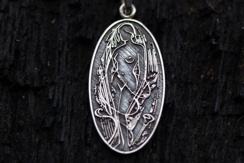 May include: Oval silver pendant featuring an engraved Art Nouveau style design. The pendant depicts a nude female figure amidst stylized floral and plant motifs. The pendant hangs from a silver chain against a dark, textured background.