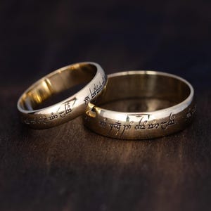 May include: Two gold-colored rings with engraved text. The rings are placed on a dark brown wooden surface. The rings are likely wedding bands or promise rings, and the engraved text adds a unique touch.