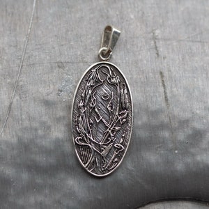 May include: Silver-toned oval pendant featuring an engraved design of a stylized figure surrounded by floral and abstract elements. The pendant has a loop for a chain and is displayed on a textured gray surface.