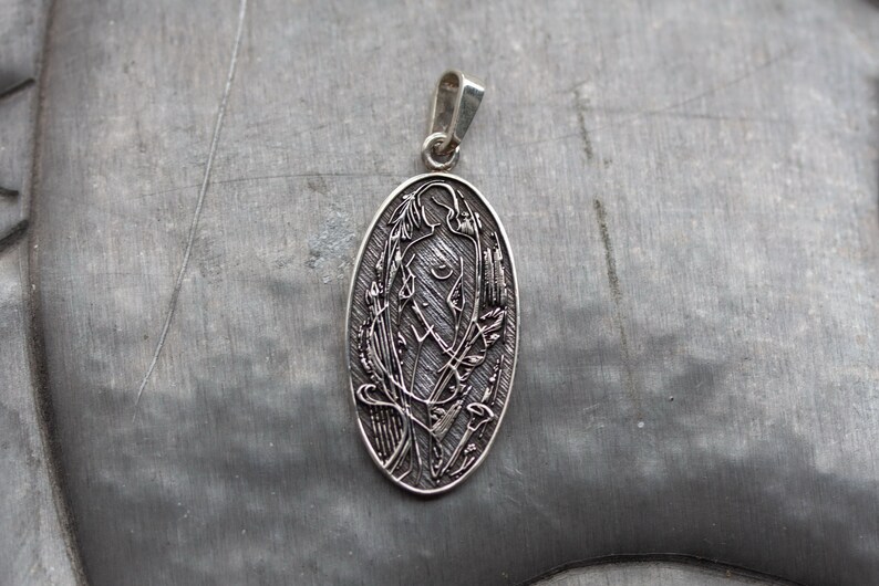 May include: Silver-toned oval pendant featuring an engraved design of a stylized figure surrounded by floral and abstract elements. The pendant has a loop for a chain and is displayed on a textured gray surface.