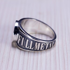 May include: A silver ring with the words "FULLMETAL" engraved on the band. The ring has a wide band and a flat top. The ring is made of a shiny, metallic material and is set against a light wood grain background.
