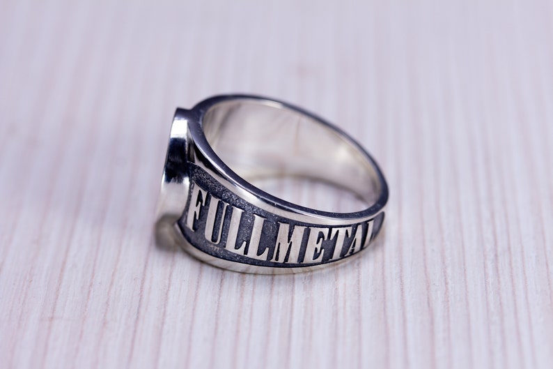 May include: A silver ring with the words "FULLMETAL" engraved on the band. The ring has a wide band and a flat top. The ring is made of a shiny, metallic material and is set against a light wood grain background.
