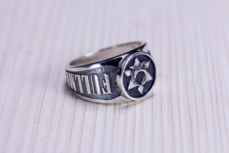 May include: A silver ring with a starburst design on a black background. The ring has the word "FULLTIME" engraved on the side. The ring is on a light wood surface.