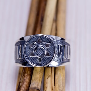 May include: A silver ring with a central design featuring a six-pointed star within a circle, surrounded by a textured background. The ring's band has raised lettering. The ring is displayed on wooden sticks.