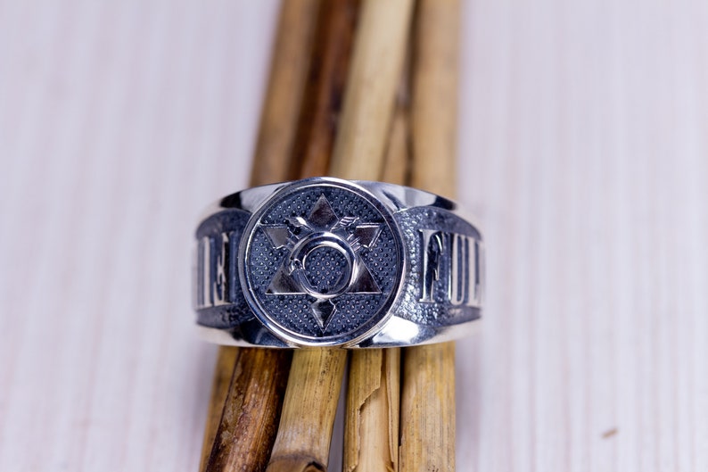 May include: A silver ring with a central design featuring a six-pointed star within a circle, surrounded by a textured background. The ring's band has raised lettering. The ring is displayed on wooden sticks.
