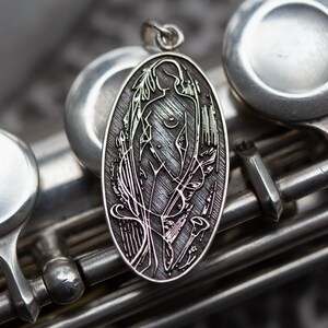 May include: Oval silver pendant featuring a stylized female figure with flowing hair and floral accents. The pendant is suspended from a small loop and is set against a backdrop of a silver musical instrument.