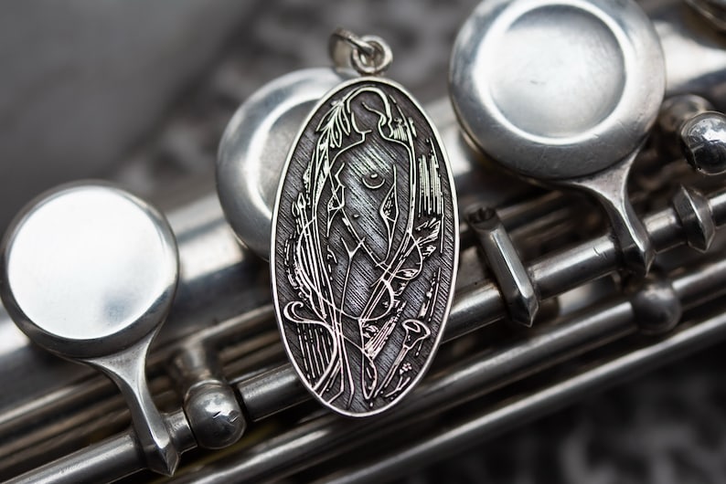 May include: Oval silver pendant featuring a stylized female figure with flowing hair and floral accents. The pendant is suspended from a small loop and is set against a backdrop of a silver musical instrument.
