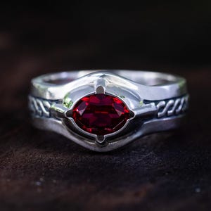 Puede incluir: Un anillo de plata con una gema roja oscura ovalada en el centro. El anillo tiene un diseño trenzado en la banda y un engaste decorativo alrededor de la piedra. El anillo está sobre un fondo de madera oscura.