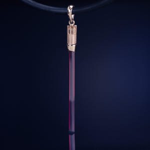 Op de afbeelding: Een goudkleurige hanger met een lange, slanke, bordeauxrode staaf die is opgehangen aan een kleine, decoratieve bovenkant. De hanger hangt aan een zwart koord tegen een donkerblauwe achtergrond. De bovenkant van de hanger heeft een textuur.