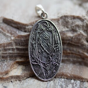 May include: Oval silver pendant featuring an Art Nouveau design of a woman with flowing hair and floral elements. The pendant is suspended from a small loop. The background is a textured, light brown stone surface.