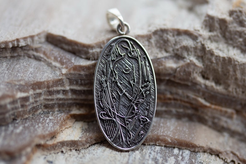 May include: Oval silver pendant featuring an Art Nouveau design of a woman with flowing hair and floral elements. The pendant is suspended from a small loop. The background is a textured, light brown stone surface.