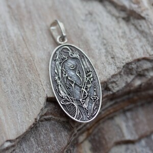 May include: Oval silver pendant with an Art Nouveau design. The pendant features a stylized female figure with floral and flowing line details. The pendant has a loop for a chain and is set against a textured background.