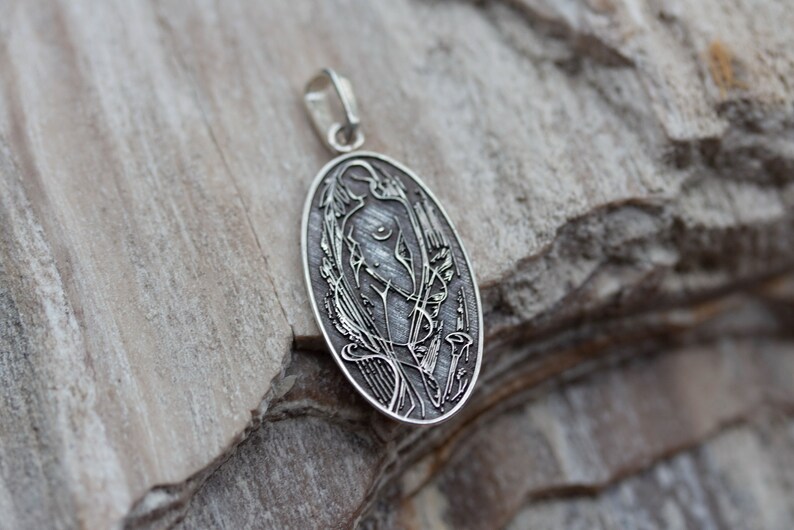 May include: Oval silver pendant with an Art Nouveau design. The pendant features a stylized female figure with floral and flowing line details. The pendant has a loop for a chain and is set against a textured background.