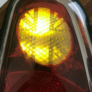 Può includere: Un primo piano di un fanale posteriore rosso con un design Union Jack giallo. Il fanale posteriore è in plastica trasparente e ha una finitura cromata.