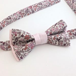 May include: A floral bow tie with a pale pink centre and trim. The bow tie features a pattern of small flowers in shades of pink, red, and purple on a white background. The bow tie is likely made of fabric.