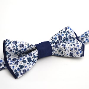 Peut inclure: Un nœud papillon blanc avec un motif floral bleu et une bordure bleu marine. Le nœud papillon est noué et prêt à être porté.