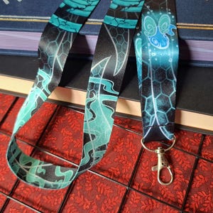 Puede incluir: Un cordón de tela con un esquema de color negro y azul verdoso que presenta un patrón repetitivo de formas abstractas azul brillante, texturas de panal y líneas onduladas, terminado con un broche de langosta de metal plateado.