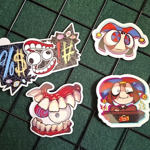 Op de afbeelding: Vier kleurrijke stickers met illustraties in cartoonstijl. Eén sticker toont een gezicht met een narrenkap. Een andere heeft een gezicht met een brede grijns en dollartekens. De andere twee stickers hebben cartoongezichten met tanden en een narrenkap.
