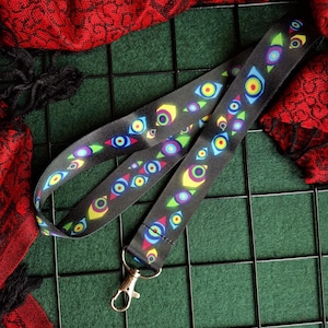 May include: A black fabric lanyard featuring a repeating pattern of stylised eyes in vibrant colours including yellow, blue, green, and purple. The lanyard includes a silver-toned metal lobster clasp at the bottom for attaching items securely.