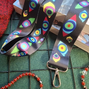 May include: Black lanyard with a repeating eye-shaped pattern in bright colors. The lanyard has a silver-colored clasp. A red beaded necklace and books are in the background.