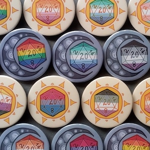 Op de afbeelding: Een verzameling van 12 ronde, kleurrijke knopen met een regenboogkleurig 20-zijdig dobbelsteendesign. Elke knop heeft een ander ontwerp met een zon- of maanmotief.