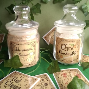 Peut inclure: Deux bocaux en verre avec des étiquettes et des couvercles. Le bocal de gauche a une étiquette qui indique "Anti-vampire Powder". Le bocal de droite a une étiquette qui indique "Ogre Dandruff".