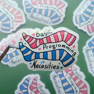 Op de afbeelding: Een sticker met een paar roze-wit gestreepte sokken en een paar blauw-wit gestreepte sokken. De tekst op de sticker luidt "Basic Programming Necessities".