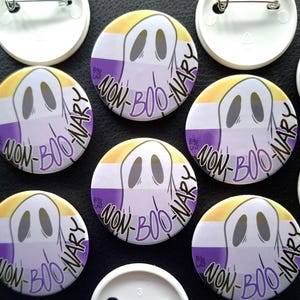 Op de afbeelding: Negen ronde pinnen met een spookontwerp en de tekst "Non-Boo-Nary" in paars, geel en wit. De pinnen liggen op een zwart oppervlak.